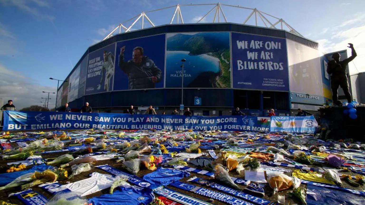  Cardiff City Stadium with blue, Bluebirds branding on the outside of it. In front of the stadium are hundreds of bunches of flowers, messages and blue Cardiff City scarves and shorts strewn on the ground. A long blue banner wrapped round the gate in front of the stadium reads: 