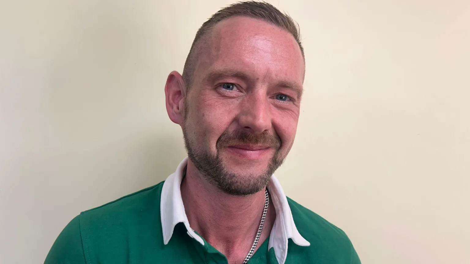 George Conway has short brown hair and a beard. He is wearing a green and white jersey and has a silver chain around his neck.