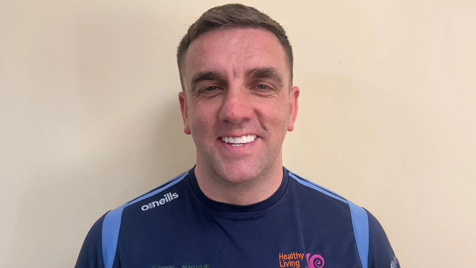 Mo Mahon has short brown hair. He is smiling and standing in front of a beige wall. He is wearing a navy and blue T-shirt.