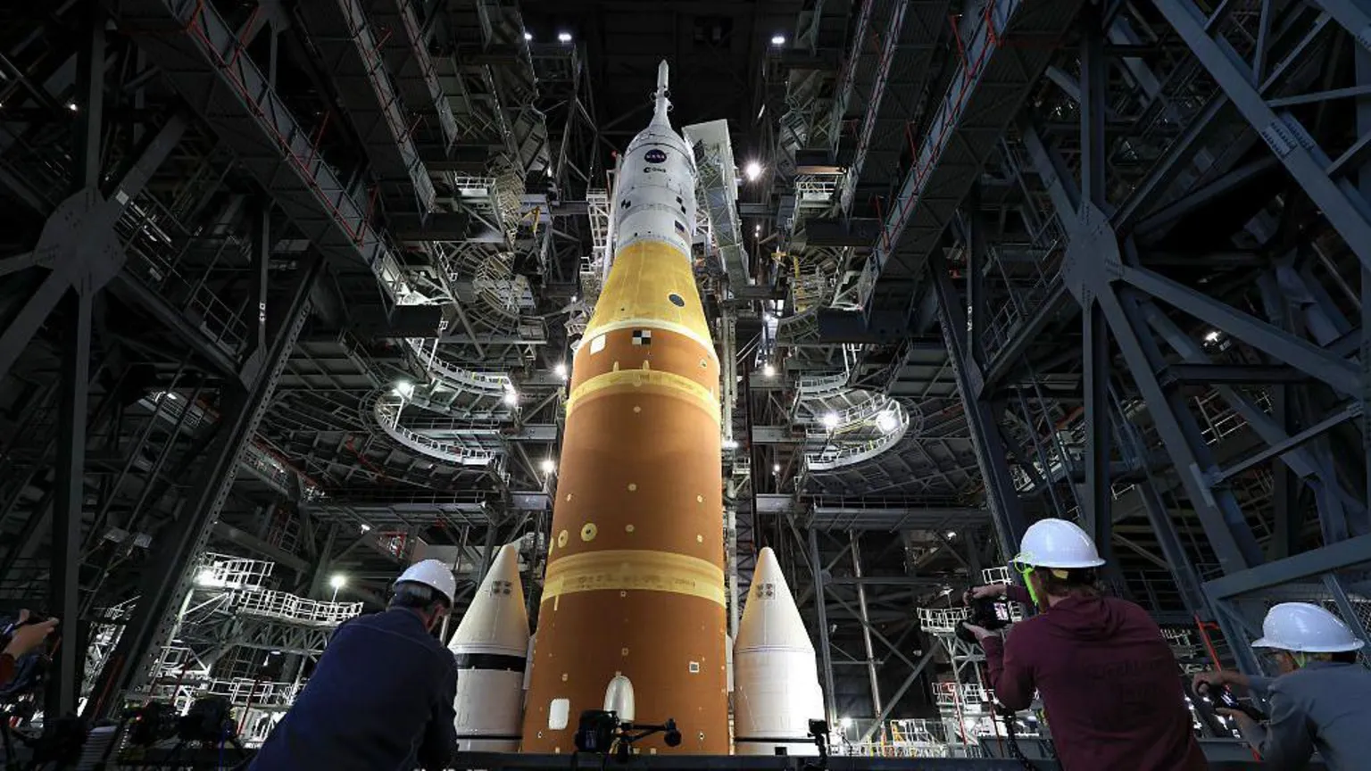 Joe Raedle/ Artemis II sits in the Vehicle Assembly Building at NASA’s Kennedy Space Center as three workers wearing white hard hats look up at the rocket