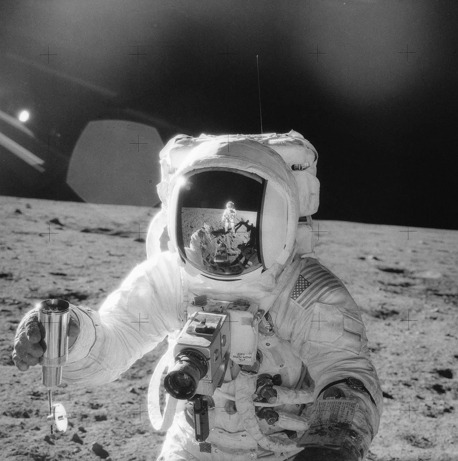 NASA Black-and-white still of Apollo 12 mission in 1969 showing one of the astronauts on the Moon’s surface holding a container of lunar soil. The other astronaut is seen reflected in his helmet.
