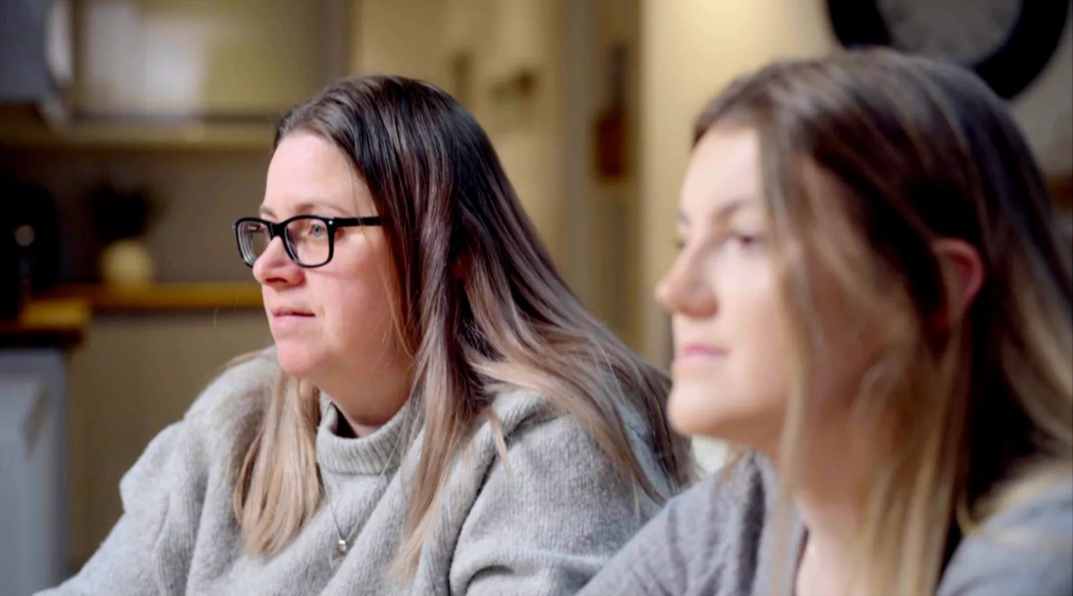 Marie, a woman with long, brown hair and dark-framed glasses, wearing a grey jumper, sits in what appears to be a family kitchen looking to the left of the camera. Next to her is Caelia, a young woman with light brown hair who is positioned nearer the camera and slightly blurred by the shallow depth of field.
