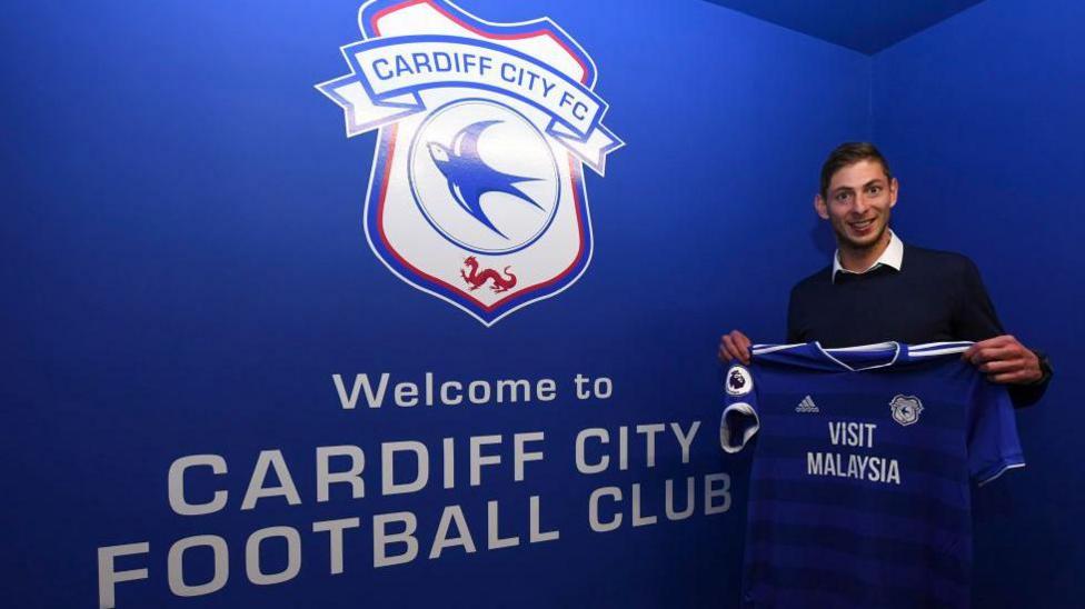 Emiliano Sala yn dal crys CPD Caerdydd ac yn sefyll o flaen logo'r clwb.