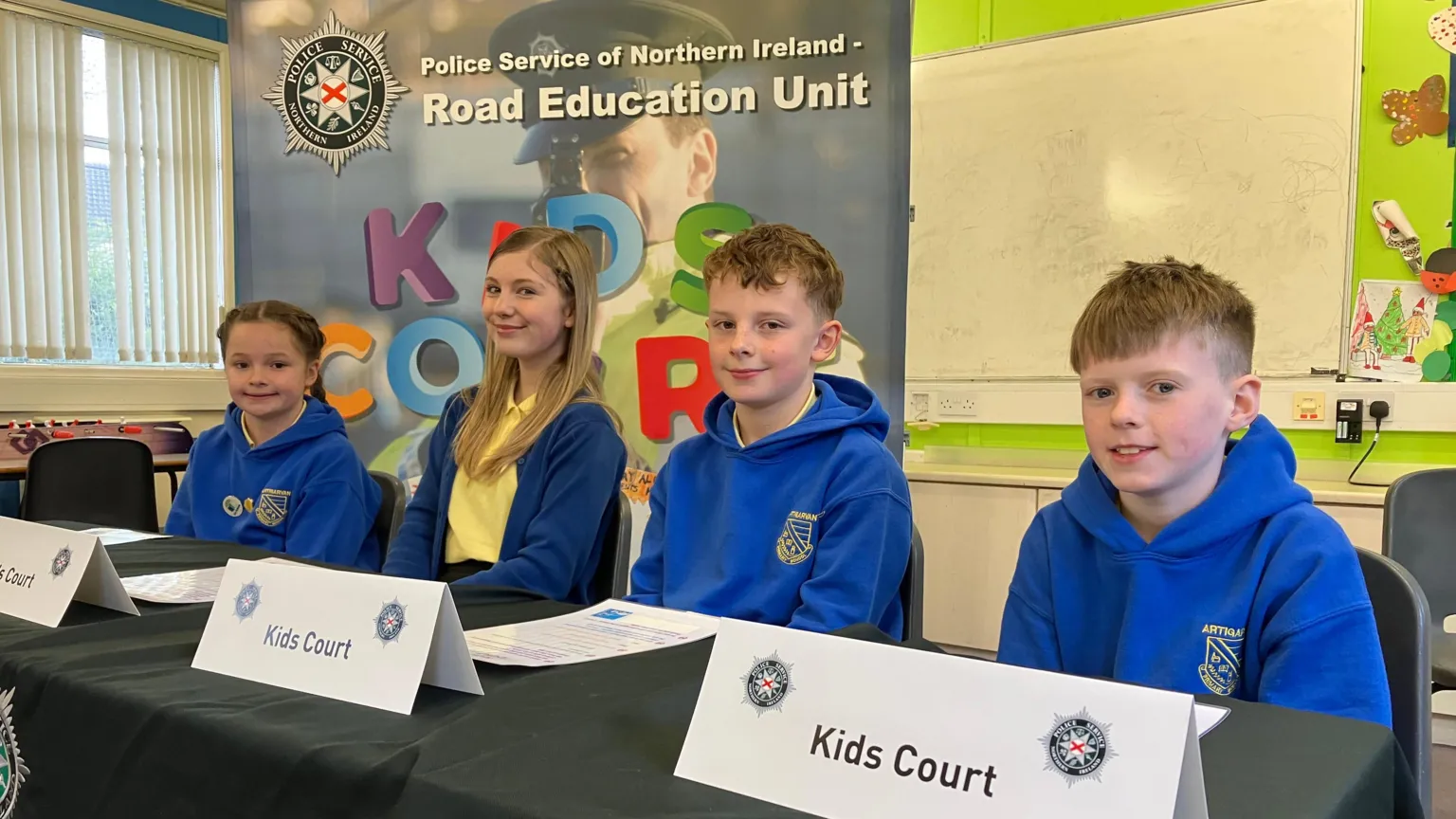 A number of schoolchildren, two girls and two boys sitting at a table in a classroom. There are several signs on the table with PSNI symbols which say 'Kids Court'. The uniform is royal blue and yellow.
