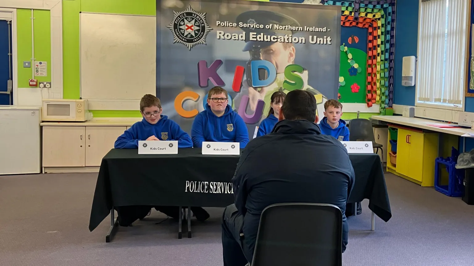 A man in grey clothes sits in a classroom facing a table where a number of schoolchildren, one girl and three boys are sitting. There are several signs on the table with PSNI symbols which say 'Kids Court'.