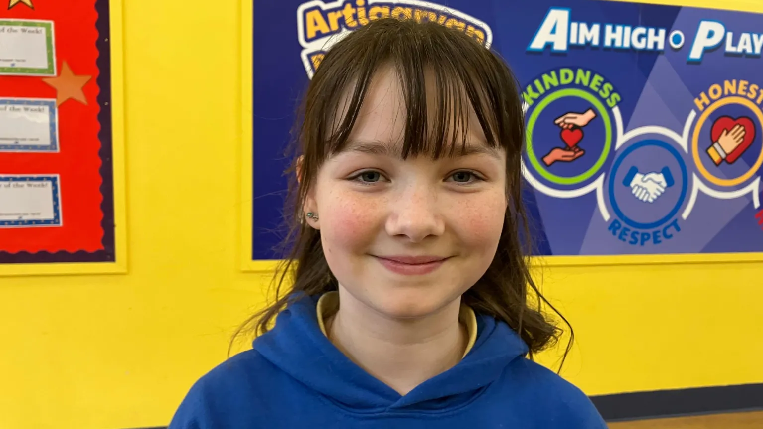 Alesha has black hair with a fringe. She has freckles and is standing in a school corridor. The wall behind her is yellow and has several posters and boards.