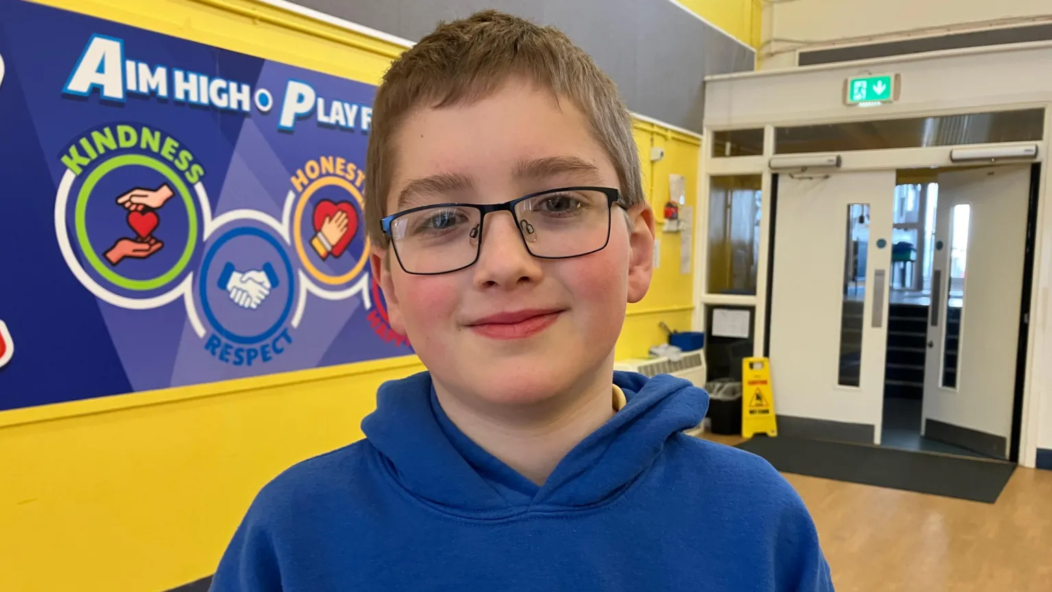 Robert has brown hair and glasses, and he is standing in a school corridor. The wall behind him is yellow and has several posters and boards.