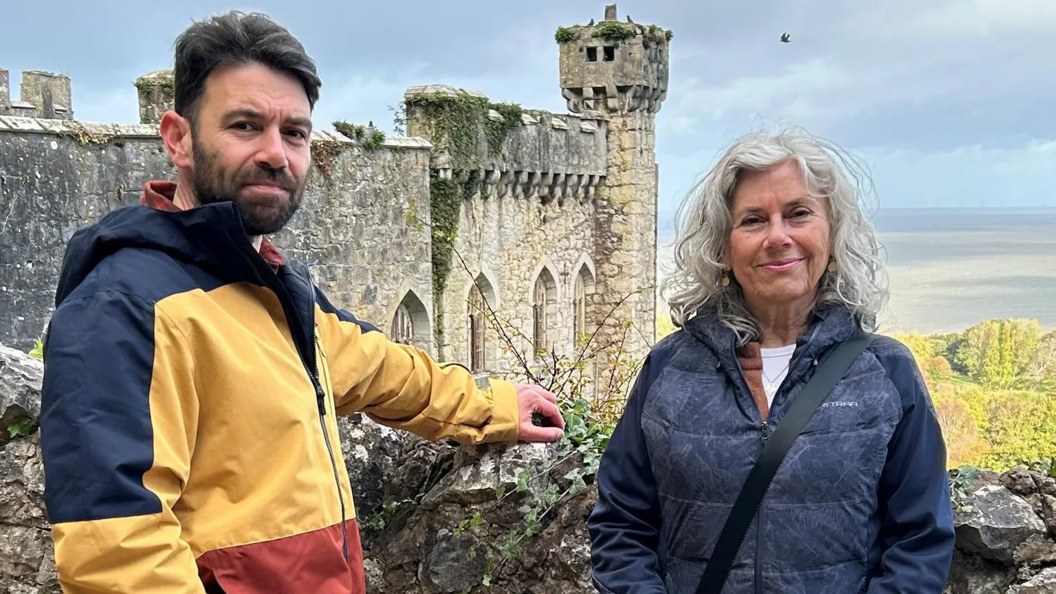 Sky Benjamin and Sandra Preiss standing against a wall with Gwrych Castle in the background. Benjamin wears a blue, white and red thick stripe coat. Sandra wears a dark blue coat.