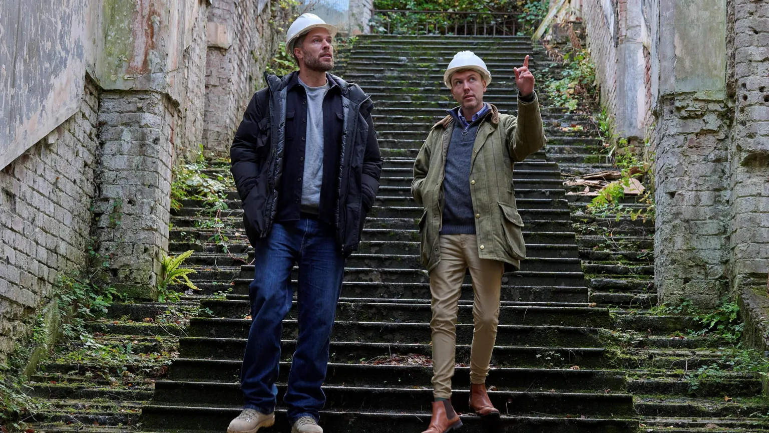 Sky Edwards and Baker walk side-by-side down stairs at the castle. Both are wearing hard hats, with Baker pointing to the sky