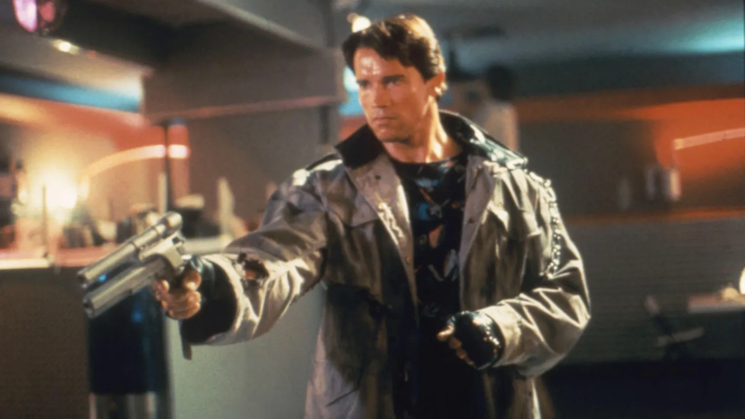 An archive picture showing Arnold Schwarzenegger on the set of Terminator. He has brown hair, is standing wearing a leather jacket and gloves pointing a scoped pistol towards the ground in front of him.