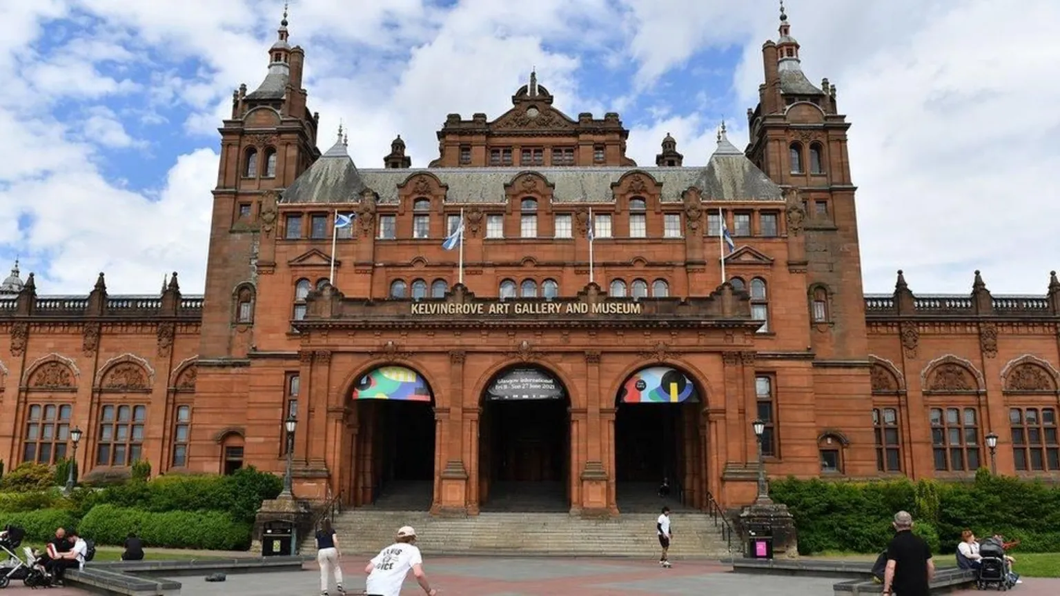  Historic red‑sandstone building with ornate towers and archways, displaying the signage ‘Kelvingrove Art Gallery and Museum,’ with people walking and sitting in the open area in front