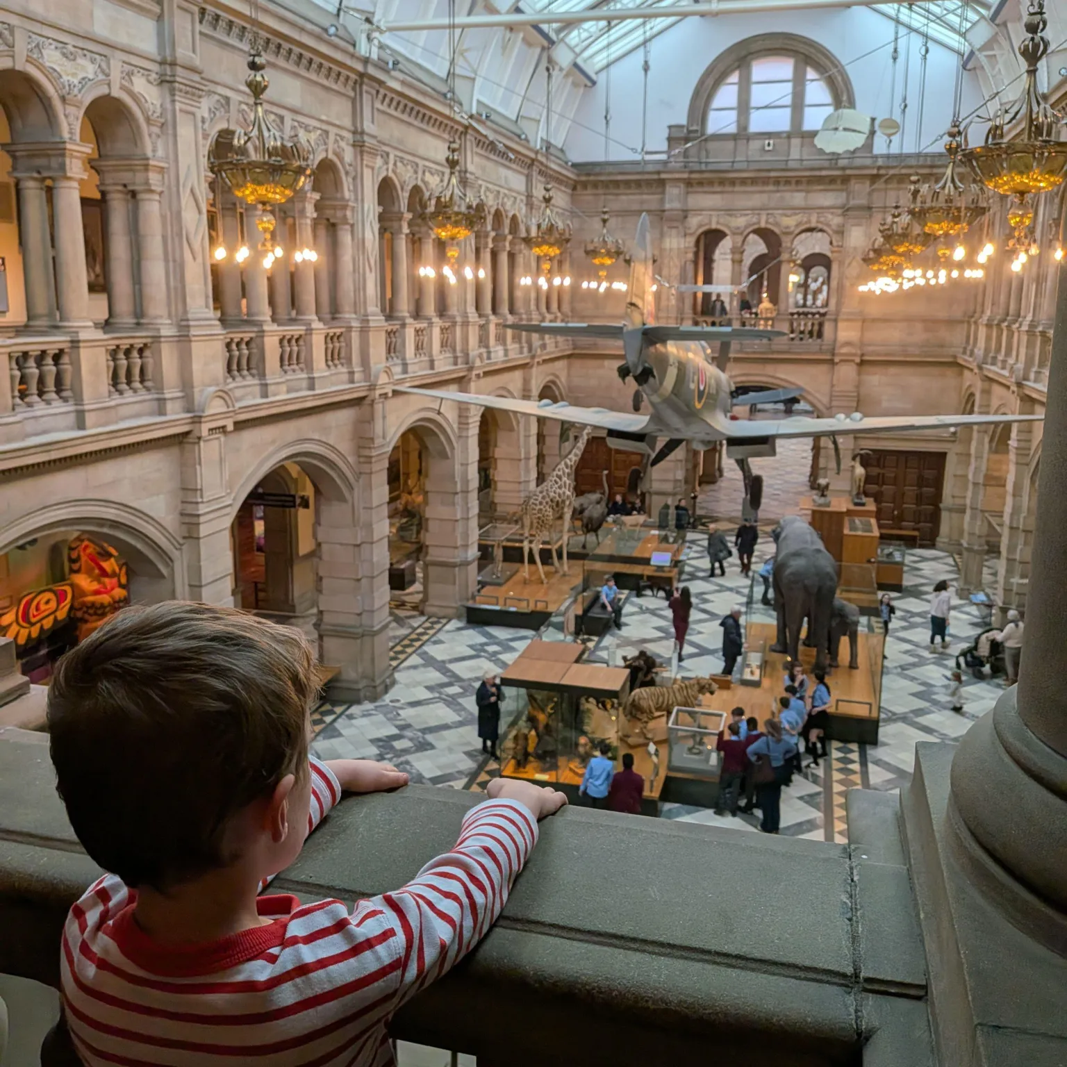 A child in a striped shirt leaning on a stone balcony inside a large museum hall with arched balconies, ornate lighting, a marble floor, and exhibits including a suspended aircraft, a giraffe model, and an elephant sculpture