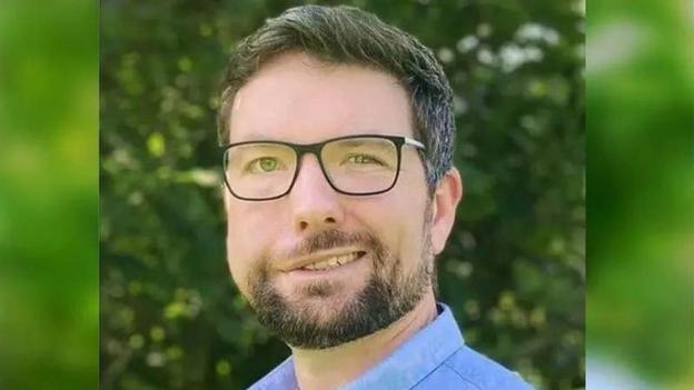 Dyn gyda gwallt brown, sbectol du a barf brown yn gwenu wrth edrych at y camera. Mae'n gwisgo crys glas.