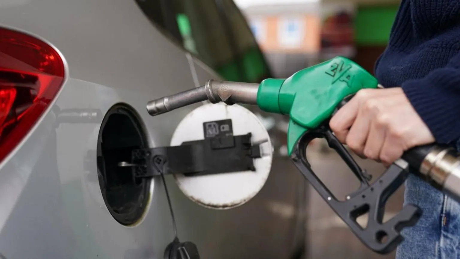  A silver car has the fuel tank lid open. A person is holding a green petrol pump ready to put fuel into the car.