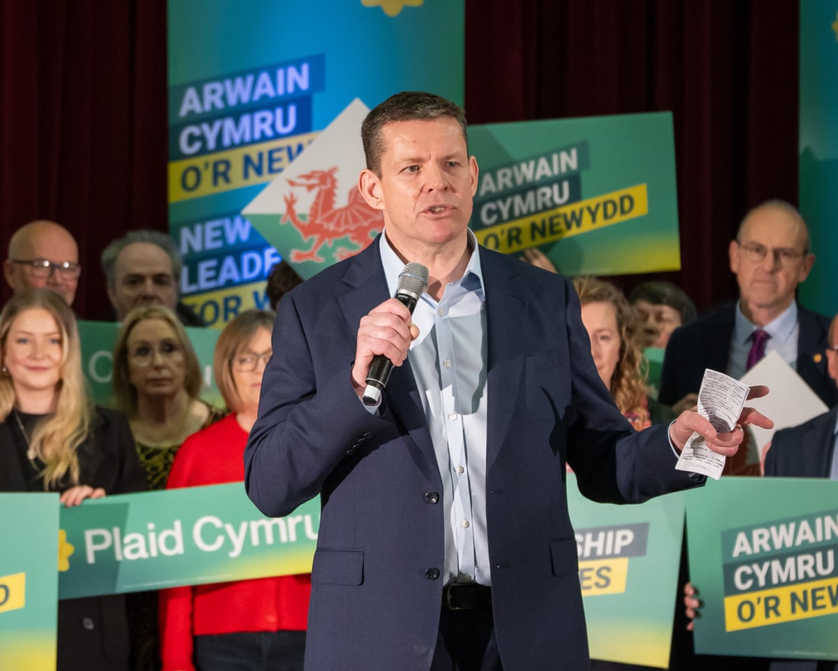 Rhun ap Iorwerth speaking, with people holding Plaid Cymru placards behind him