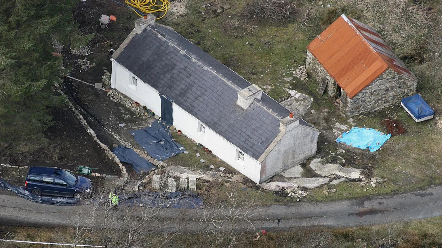 An aerial image of a white washed house in a remote location. S shed is behind the house and trees are around it. A police officer is standing nearby.