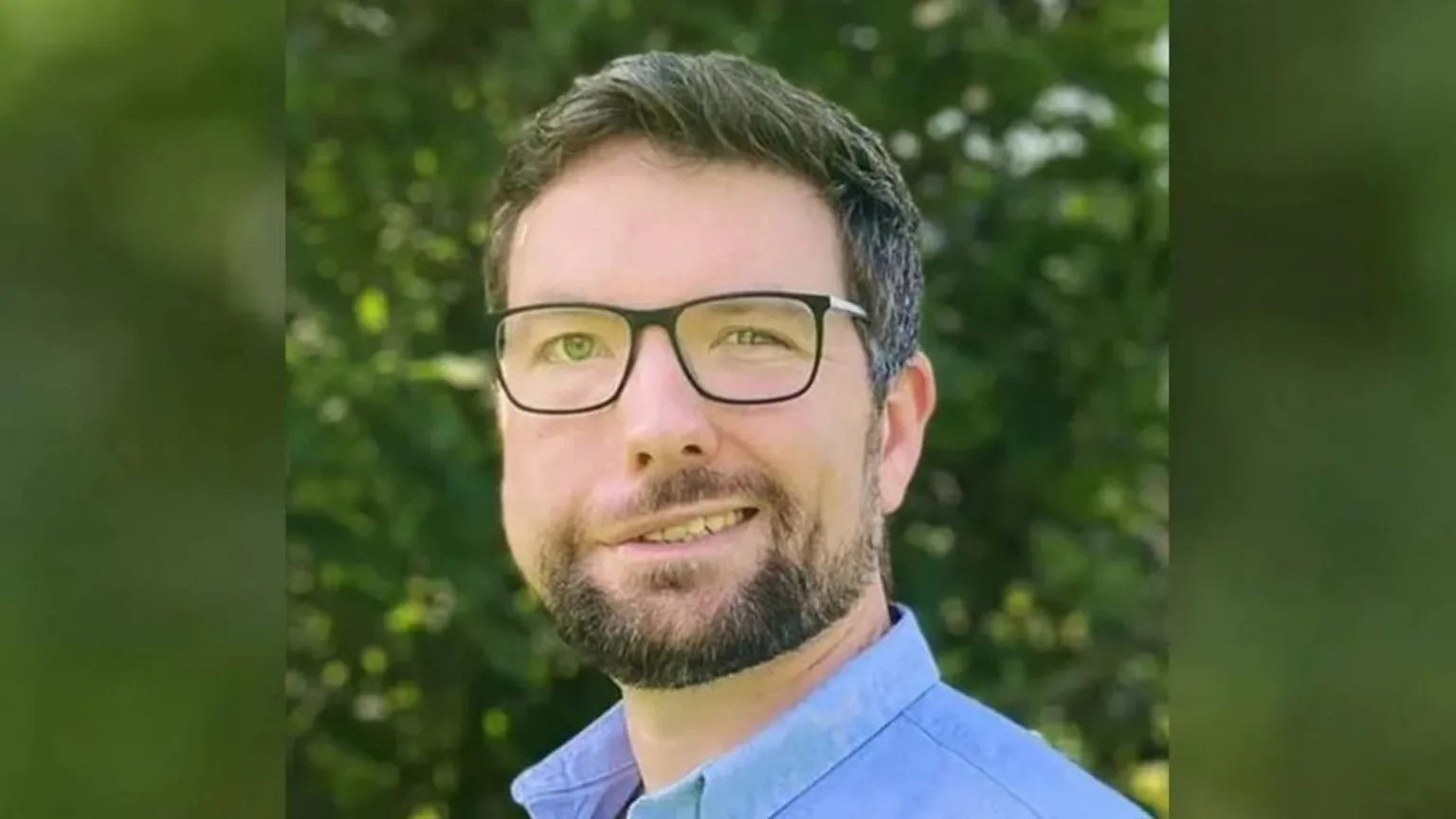 Family photo Aaron Jones looking at the camera. He is wearing a blue shirt and dark-rimmed glasses and dark hair. He has a short trimmed beard and moustache
