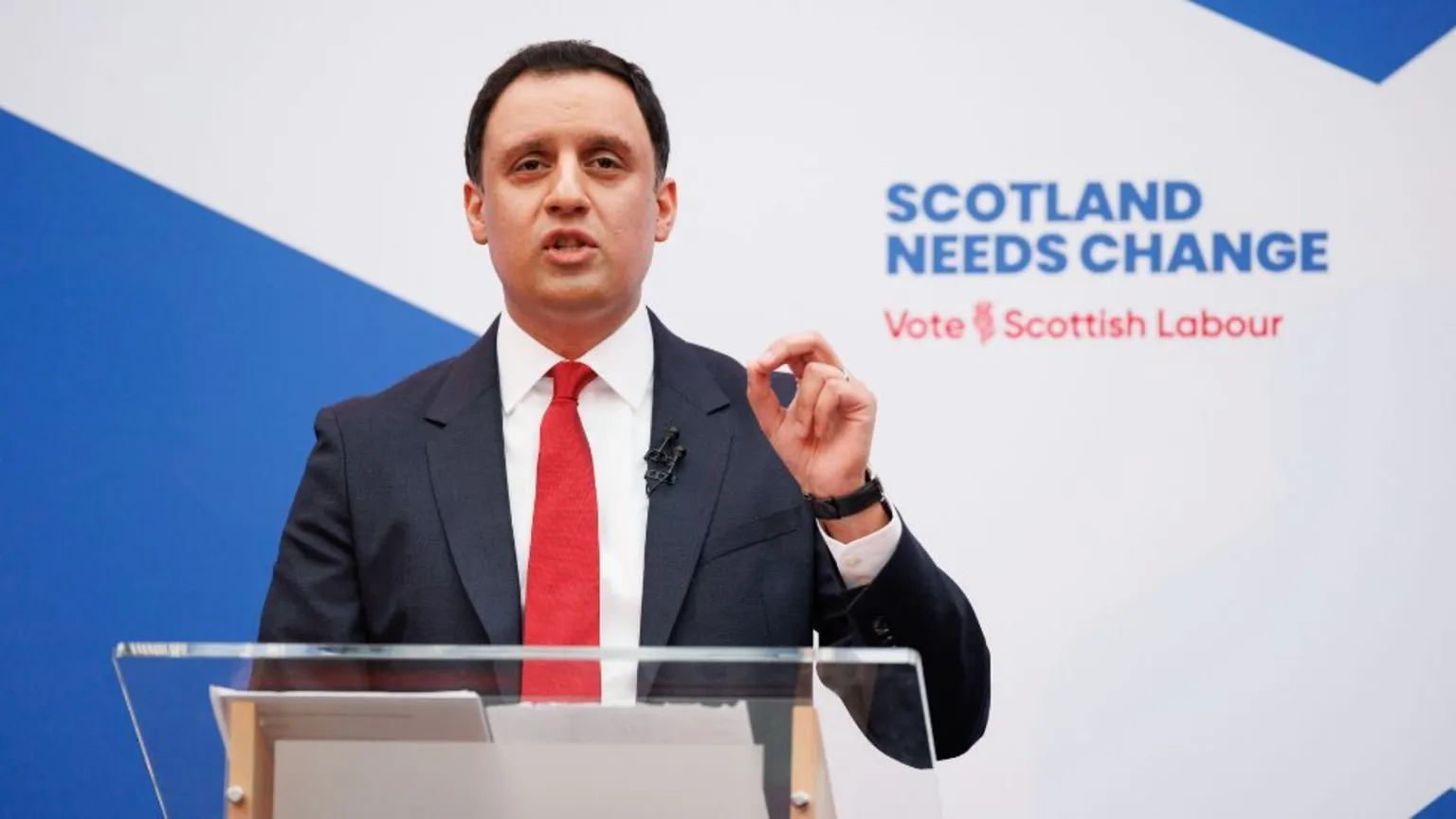  Anas Sarwar, who has short black hair, speaks at a podium while wearing a business suit and red tie. There is Labour branding behind him. 