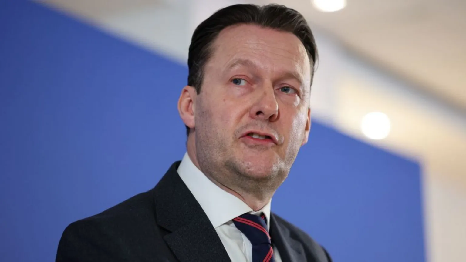  Russell Findlay, who has dark hair swept back, speaks in front of a blue background. He is wearing a business suit 