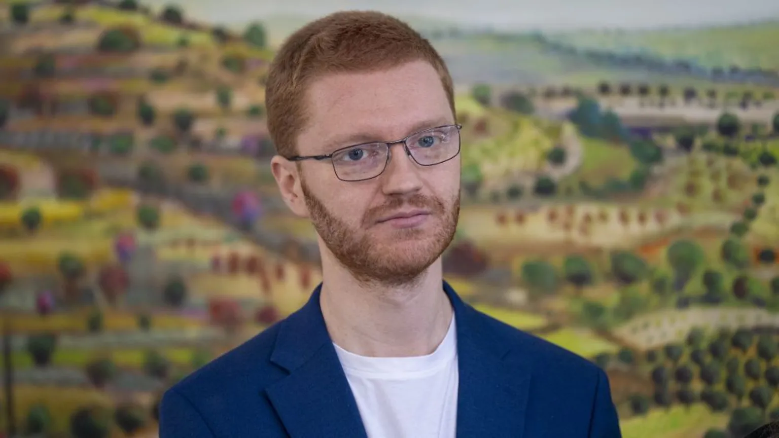  Ross Greer, who has short ginger hair and glasses, in a close-up shot. There is a picture of green fields behind him. 