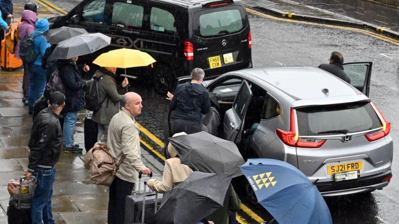 A taxi and a private hire car dropping people with umbrellas off in Edinburgh on a rainy day.