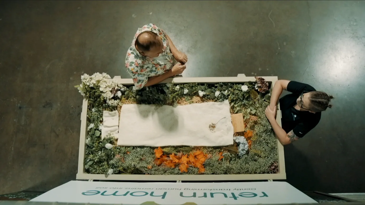 Ffilmiau Twm Twm Overhead shot, showing a human composting vessel filled with flowers and organic matter, with a small white sheet laid on the top. In front of the vessel is Kristopher Hughes, wearing a colourful shirt, and opposite at the end of the vessel is one of Return Home's staff members, a women dressed in a black t-shirt with her hair tied back