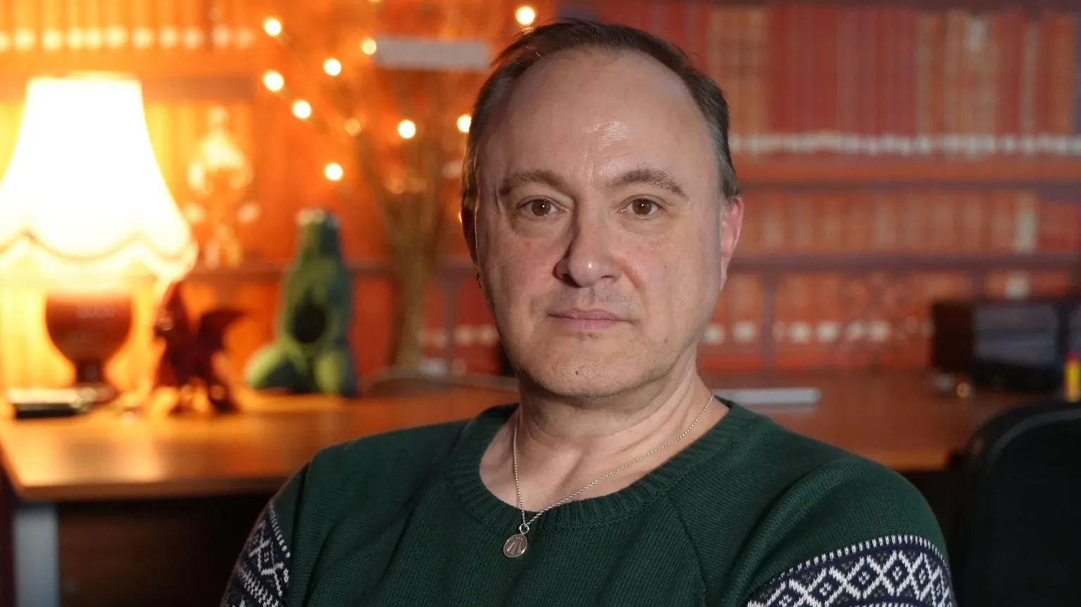 Kristoffer Hughes seated indoors at the office of the Anglesey Druid Order, wearing a green patterned sweater, wearing a silver druidic Awen symbol on a chain. It is a warmly lit room in the background featuring a desk, lamp, mock bookshelves, and decorative lights