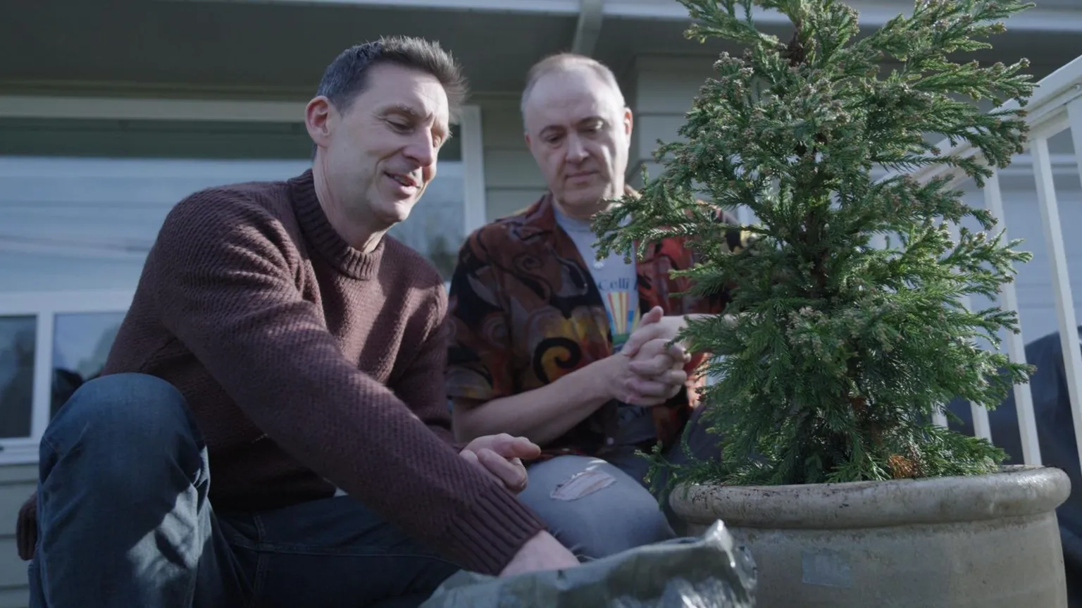 Ffilmiau Twm Twm Two men are crouched down in front of a potted evergreen shrib, one wearing a brown knitted jumper has his hand in a bag of compost, while the other man, wearing a colourful shirt and t-shirt looks on