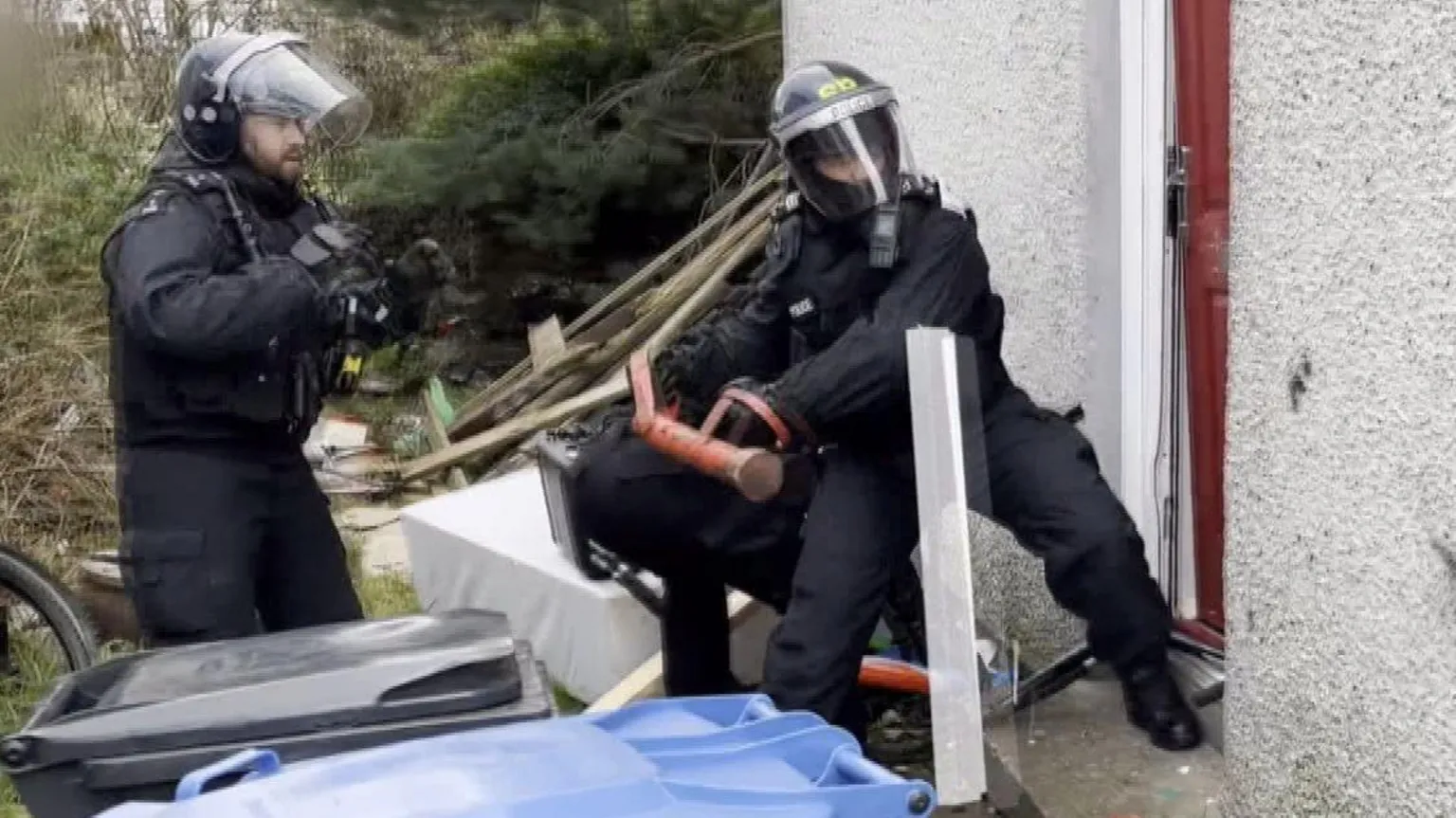 Two police officers breaking into a run-down property with rubbish covering the back garden. One officer uses a battering ram and is smashing in the property door. Both are wearing protective head gear and bullet proof vests.