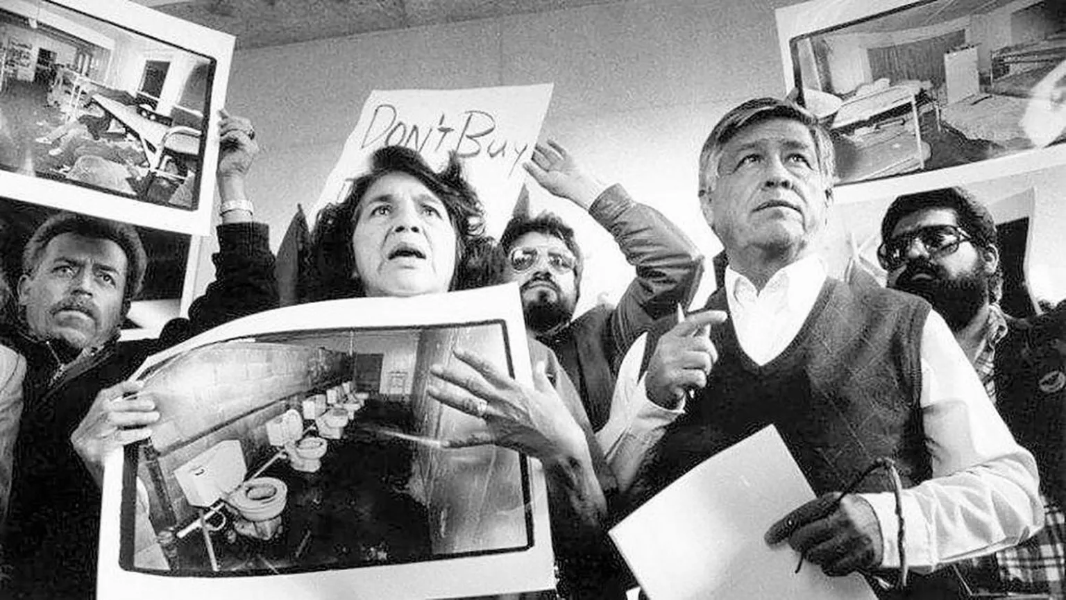 A black and white photo of Huerta and Chavez in the centre, holding up images of poor living conditions for workers, surrounded by other protestors
