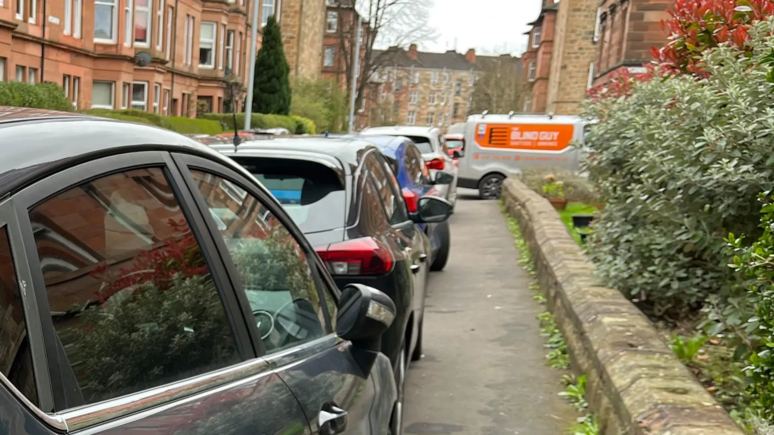 A row of parked cars lines a narrow residential street beside red‑sandstone tenement buildings. A low stone wall runs along the right side with dense shrubs and plants behind it. An orange‑and‑white van is visible farther down the street near more tenement buildings under an overcast sky.