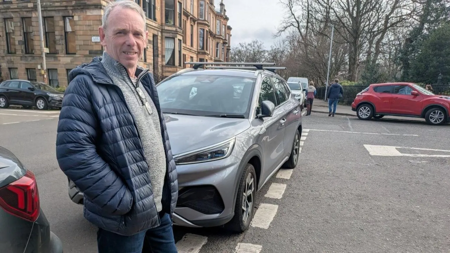 Andrew Downie wearing a padded jacket and standing beside a grey SUV parked on a residential street. Additional parked cars are visible along the road, with red‑sandstone buildings on the left and trees in the background.