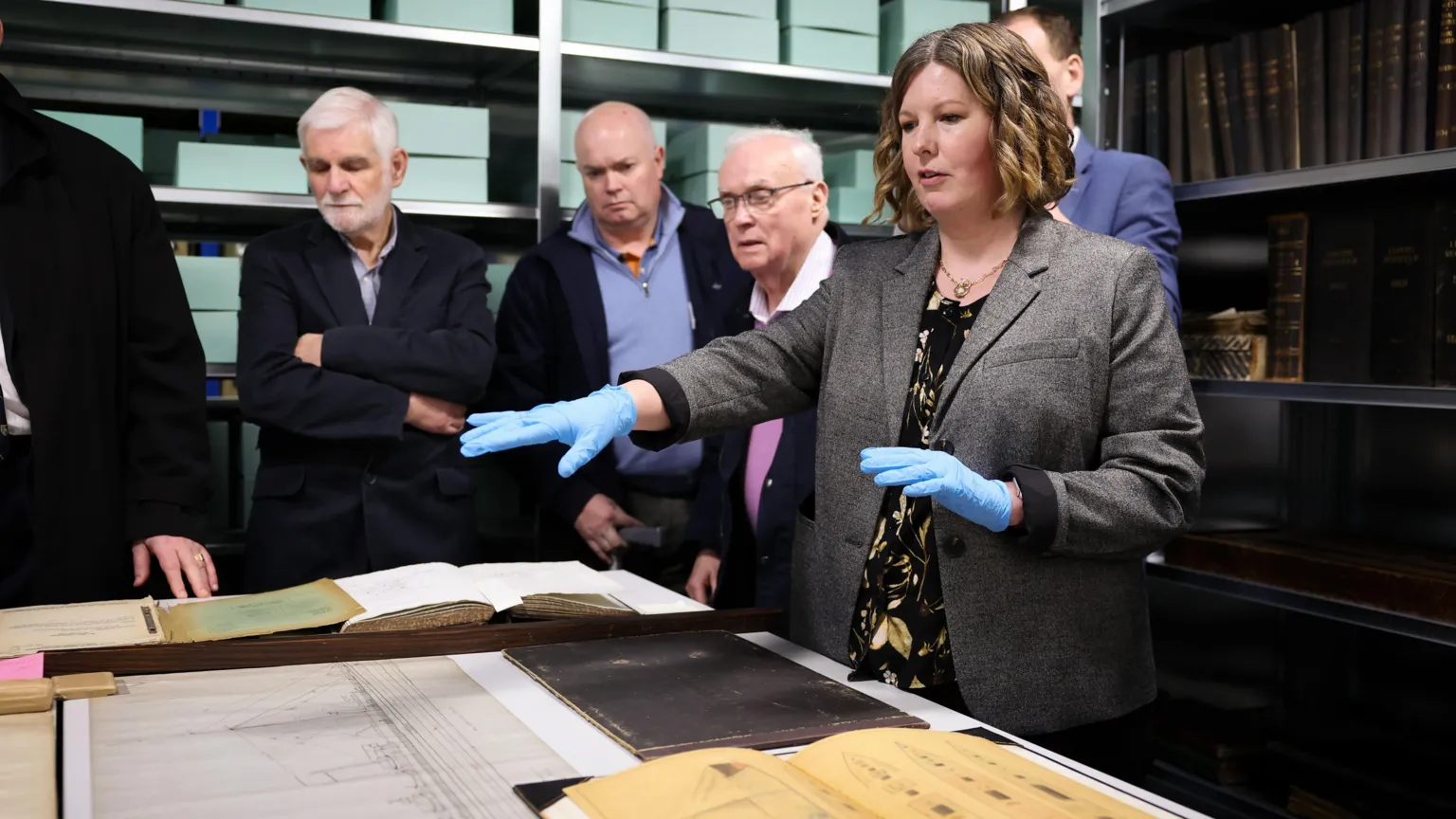 National Museums Northern Ireland Six people stand looking at a table. The table has a number of documents on it, many of which appear old. The woman in the forefront is wearing bright blue gloves. She has shoulder-length curly brown hair smiles and is wearing a grey blazer. 