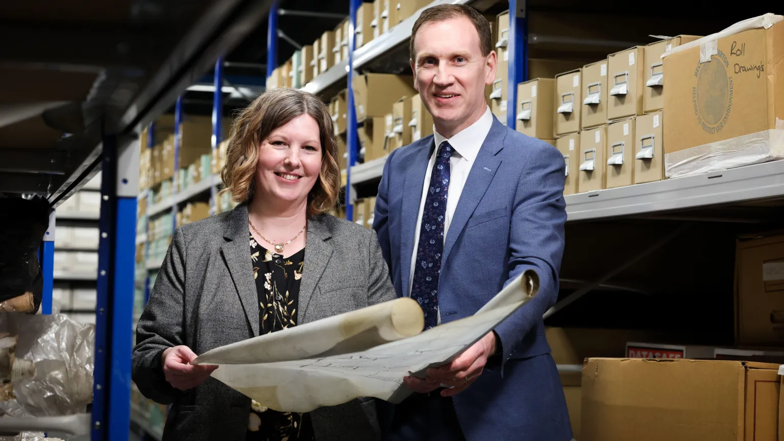 National Museums Northern Ireland A woman and a man stand together, smiling at the camera. The woman, who is stood on the left, has shoulder-length curly brown hair and is wearing a grey blazer. The man is wearing a white shirt with a blue suit-jacket. They are holding a large document. 