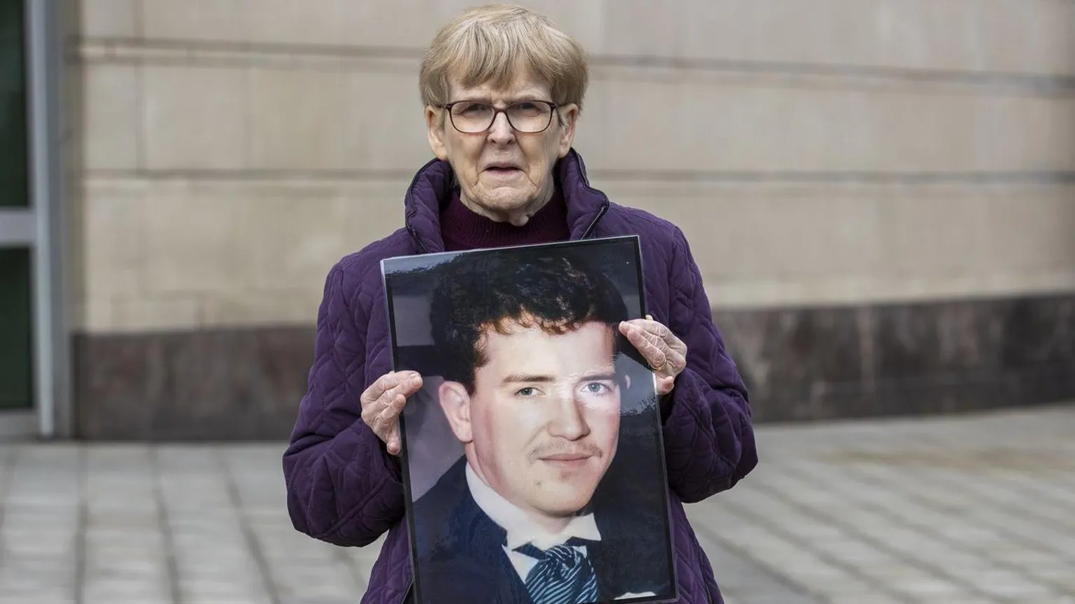  Christina McCusker has short grey hair and glasses and is wearing a purple coat. She is holding up a picture of her son Fergal who has reddish brown hair and a light moustache nad is wearing a wedding suit 
