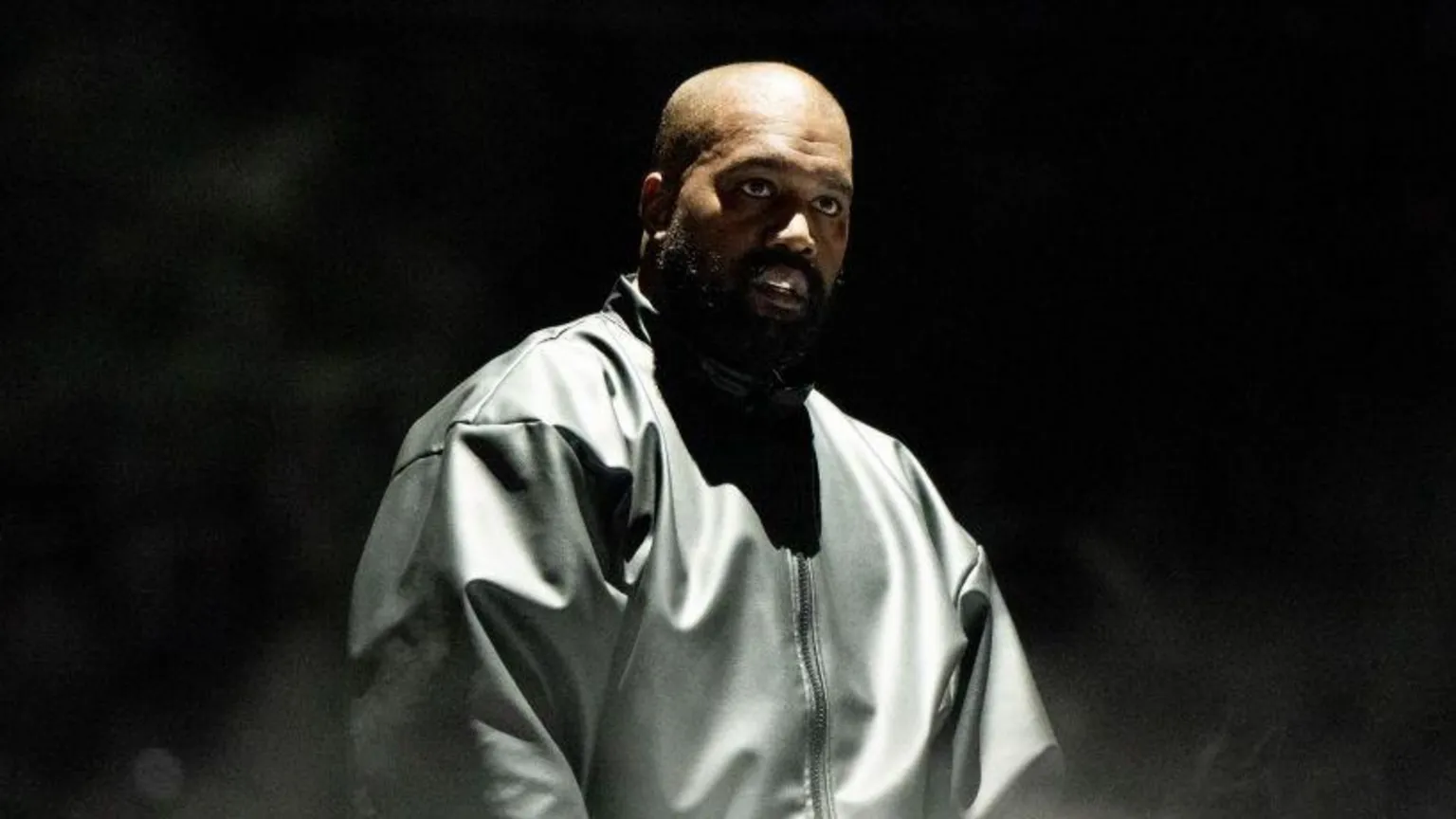  Kanye West wears a black leather jacket and has a beam of light shining down upon him while he stands in front of a black background.