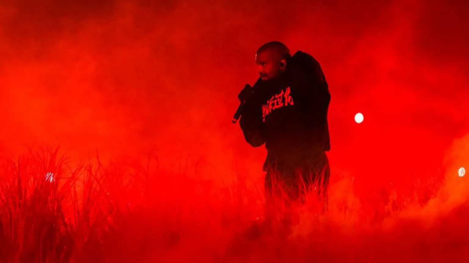  Kanye West is performing on stage. He is hunched over with a mic in one hand, which he is holding up to his face. He is wearing a black hoodie with writing in a different language on it and black trousers. He is surrounded by straw sprouting up from the ground and clouds of smoke. There are bright red lights in the background.
