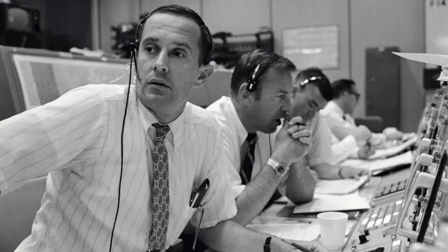 NASA Black and white image of Charlie Duke, along with Jim Lovell and Fred Haise in Nasa Mission Control, during the Apollo 11 mission. They are all dressed in shirts and ties and wear headsets, as they sit in a long line in front of control panels and display screens.