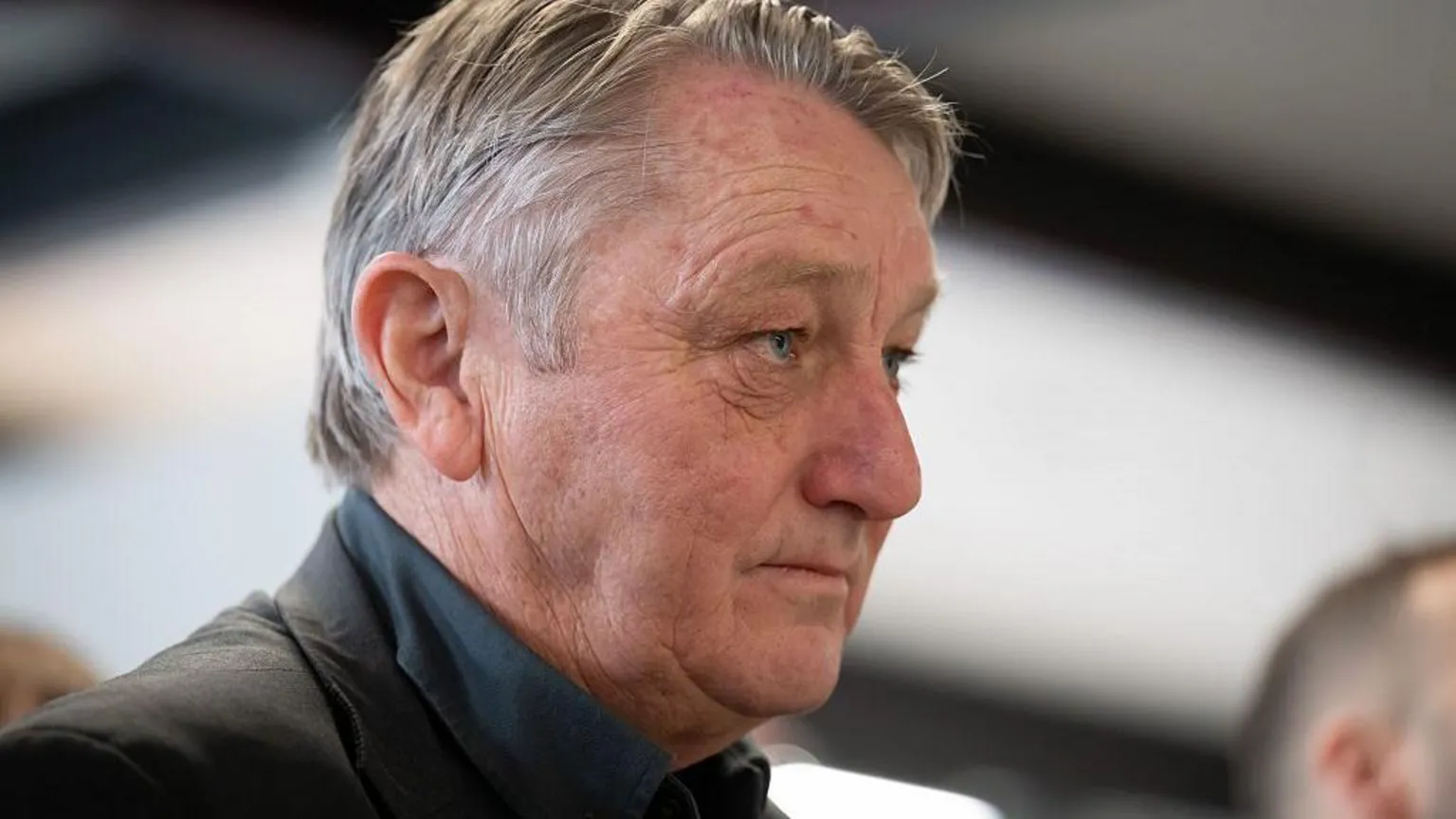 Matthew Horwood/ Anthony Slaughter with a dark suit and dark green shirt looking to the right of the picture. He has grey hair.