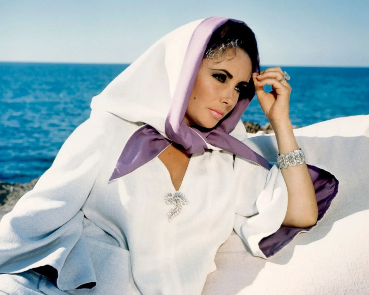 Elizabeth Taylor in a white top and white and purple scarf with the sea in the background.