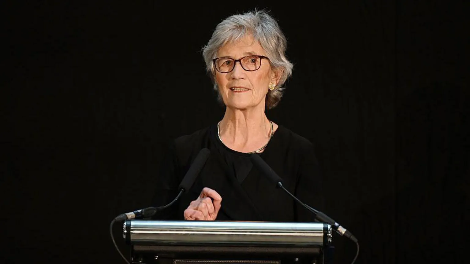 A woman with short grey hair and glasses stands at a lectern. She is wearing a black top, and the background is black. 