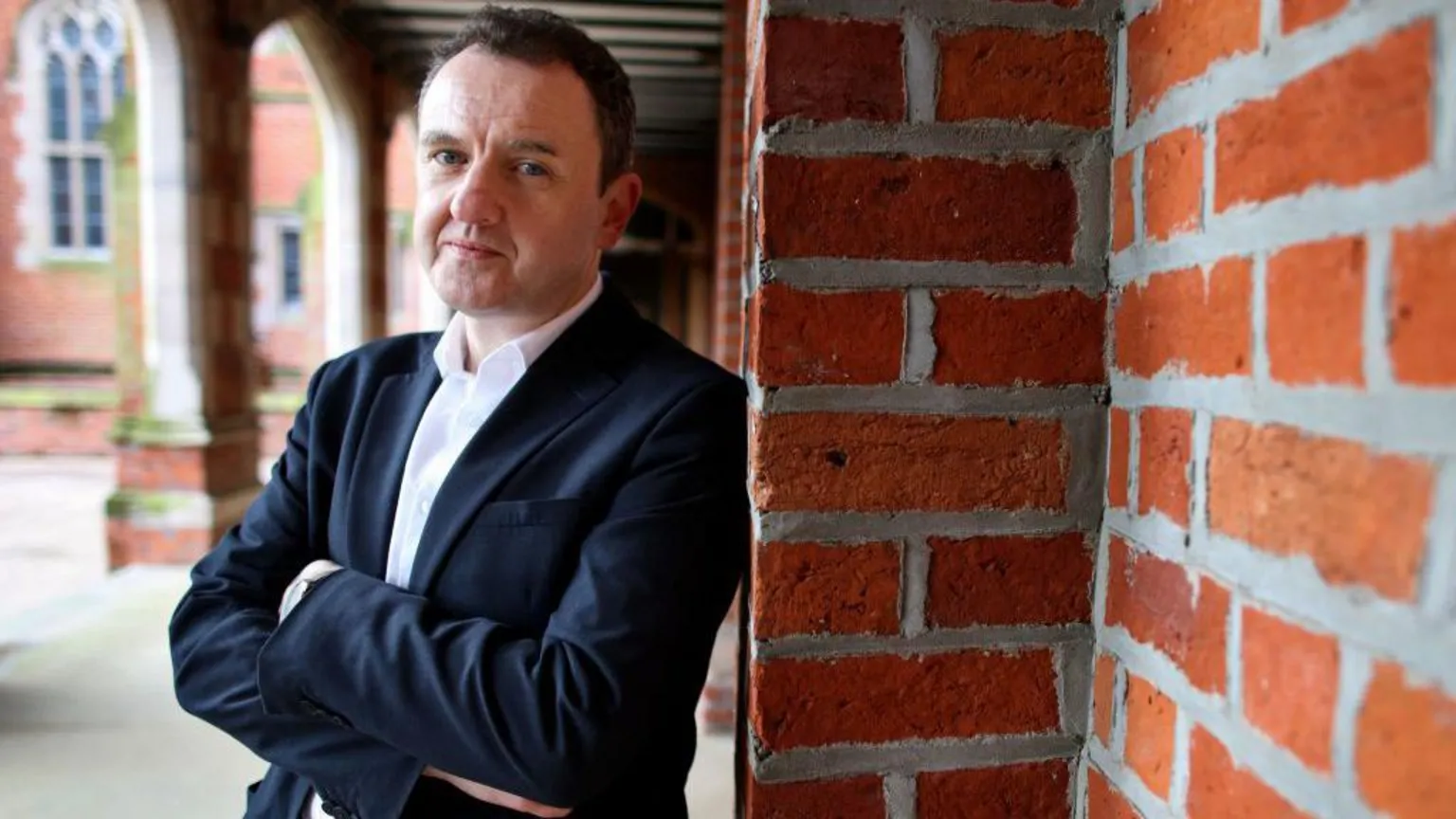  A man with a white shirt and a dark suit jacket leans against a red-brick wall. He has his arms crossed and he is looking at the camera. 