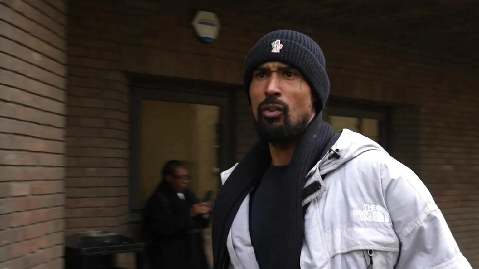 Ashley Warren walks outside the bricked frontage of Chelmsford Crown Court, wearing a white coat and dark-coloured beanie hat. He is mouthing something towards people behind the camera.