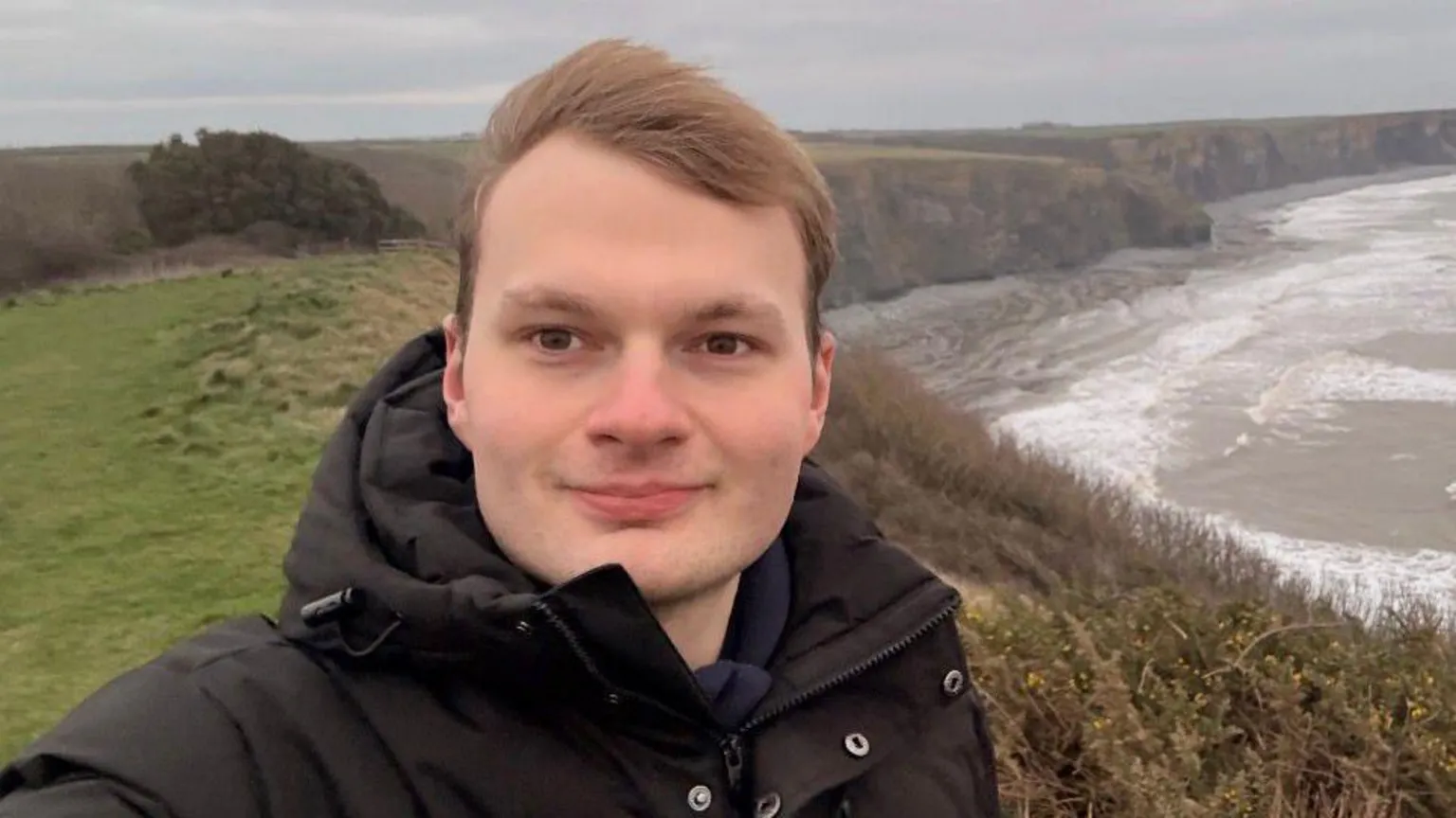 Owain Clatworthy A selfie picture of Owain Clatworthy, stood on a coast line with the cliff face twisting into the background