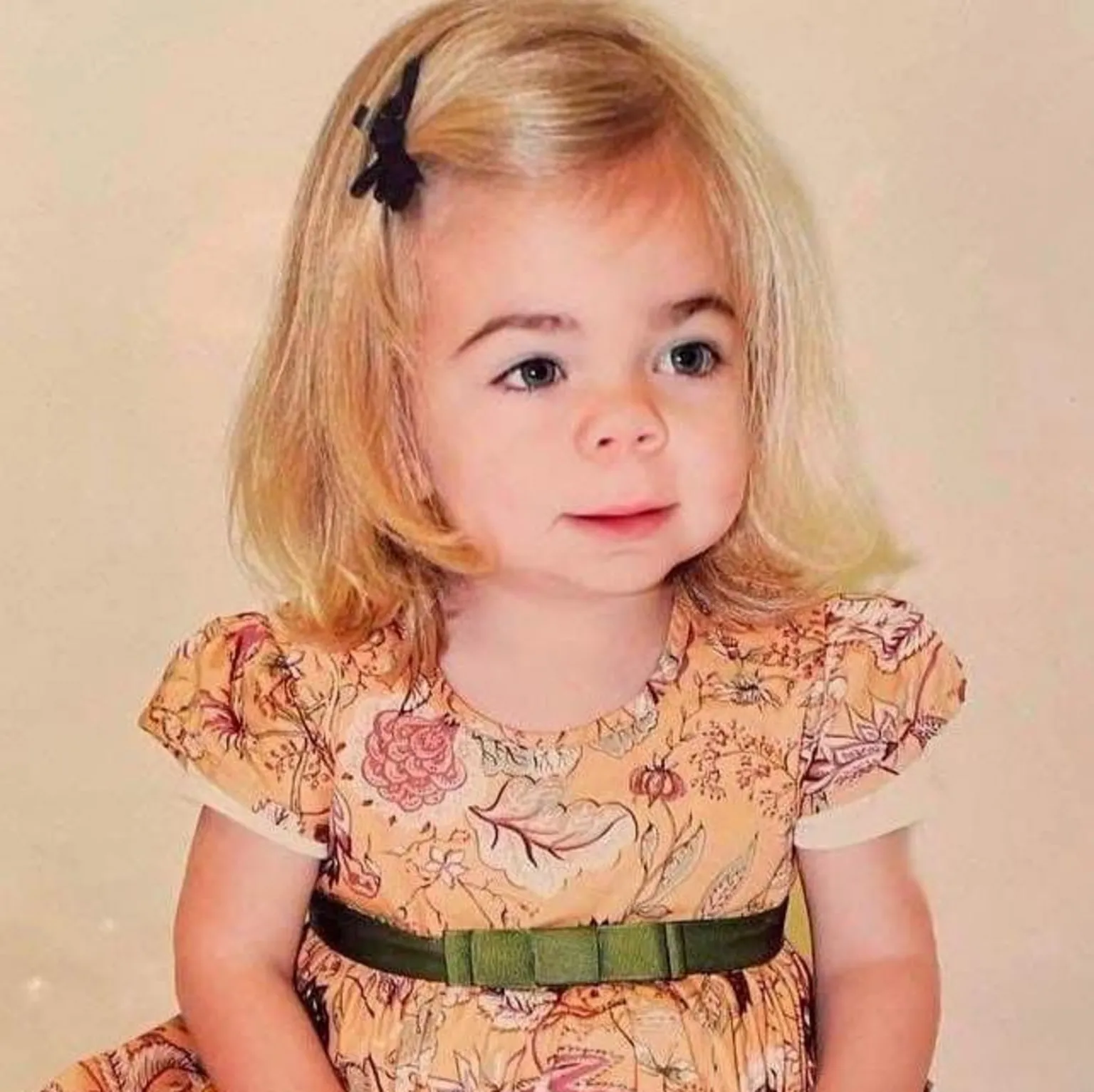 Darren Scott Sophia as a very young child. She has shoulder-length, blonde hair with a black bow clipping it away from her face. She is looking off camera and is wearing an autumnal brown dress with short sleeves and a green bow on a ribbon around it