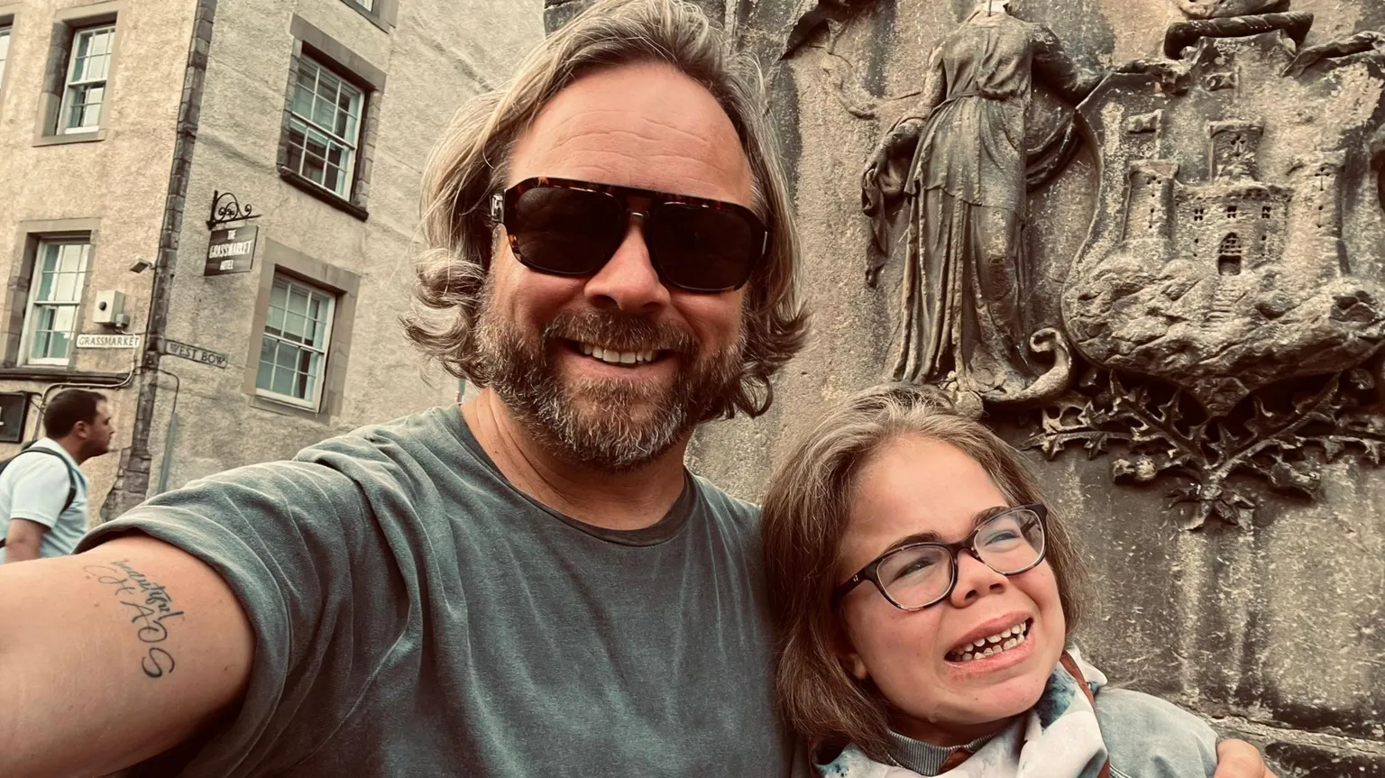 Darren Scott The image shows Darren and Sophia Scott in a selfie. He has longish blond hair and a beard and is wearing sunglasses and a grey T‑shirt. His arm is around Sophia who is wearing glasses and a scarf. They are both smiling. Behind them is a weathered stone monument or sculpture featuring carved figures and what appears to be a crest or coat of arms.