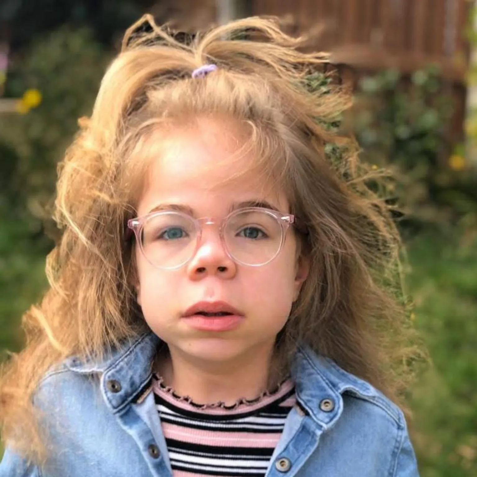 Darren Scott Sophia Scott, a young girl, is looking at the camera. She has long, blonde hair, with some of it party tied at the top with a bobble. She is wearing pale pink-rimmed glasses, a denim shirt or jacket with a pink, grey, black and white striped top underneath.