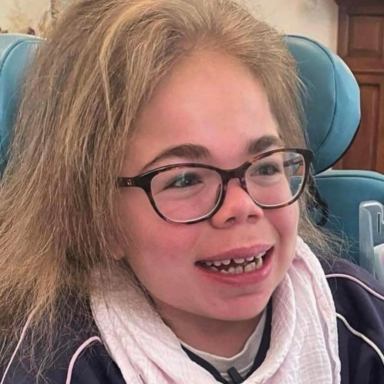 Darren Scott Sophia Scott is smiling broadly, looking off camera. She has long, blonde hair and brown-rimmed glasses. She is wearing a blue top with pink trim and a pale pink cotton scarf around her neck.