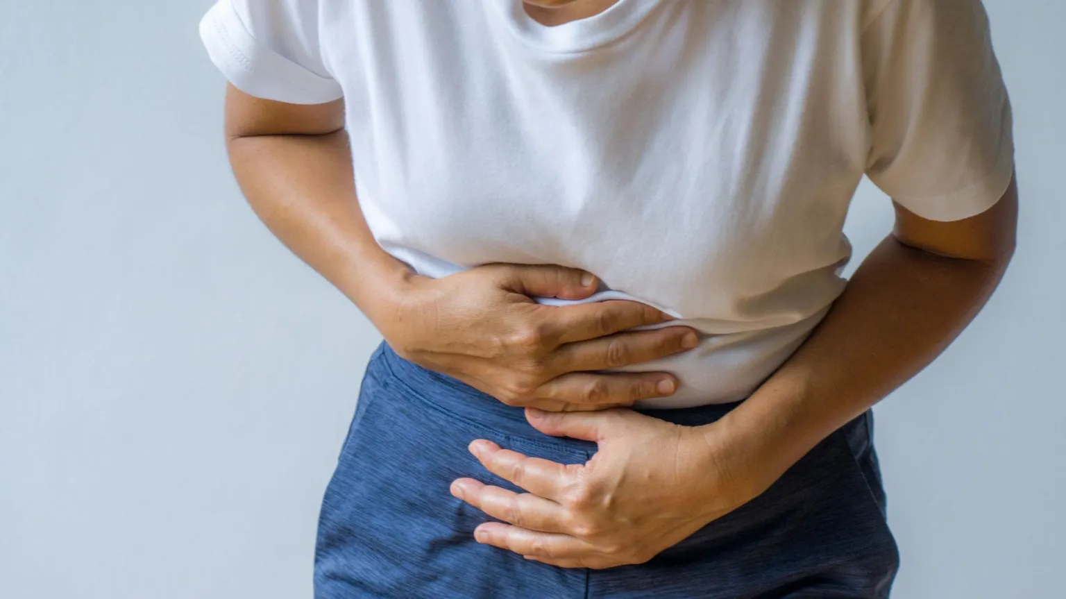A person is holding their tummy with both hands. They have a white t-shirt and navy trousers on. The background is white.