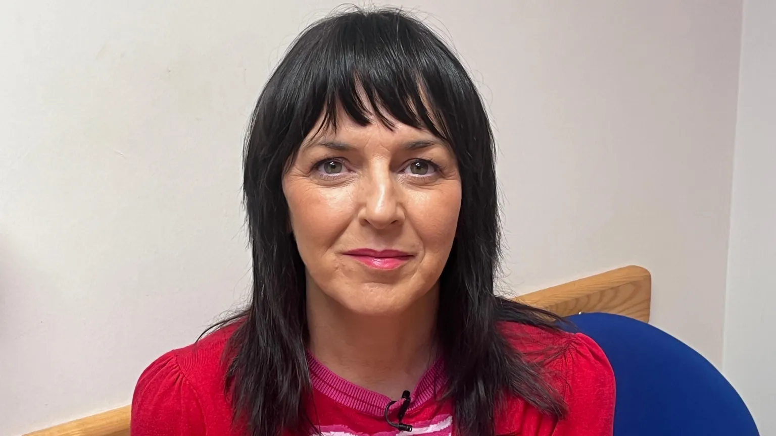 Marie McGrath is sitting on a blue chair. She has long black hair and is wearing a red top.